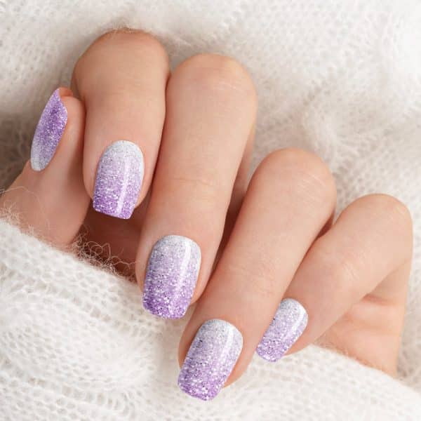 Close-Up Of A Hand With Short Nails, Each Featuring A Gradient Of White And Lavender Glitter Nail Polish.