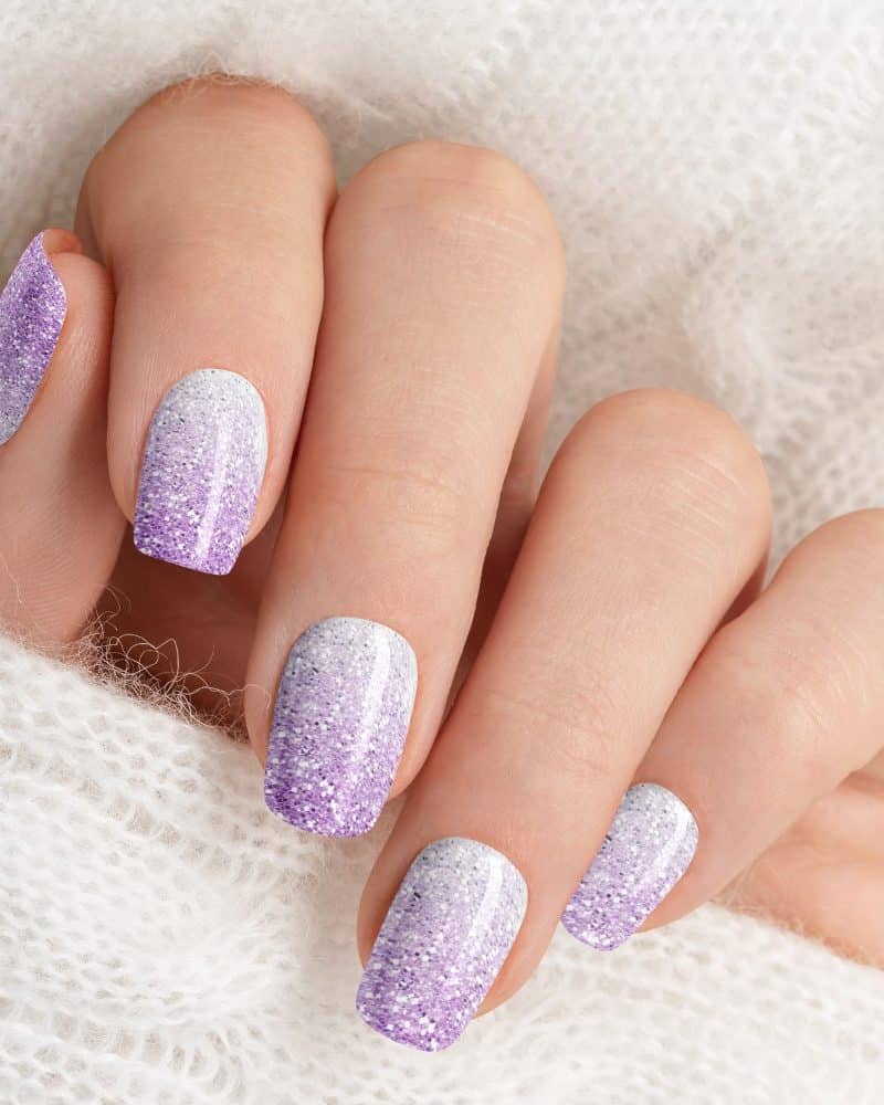 Close-up of a hand with short nails, each featuring a gradient of white and lavender glitter nail polish.