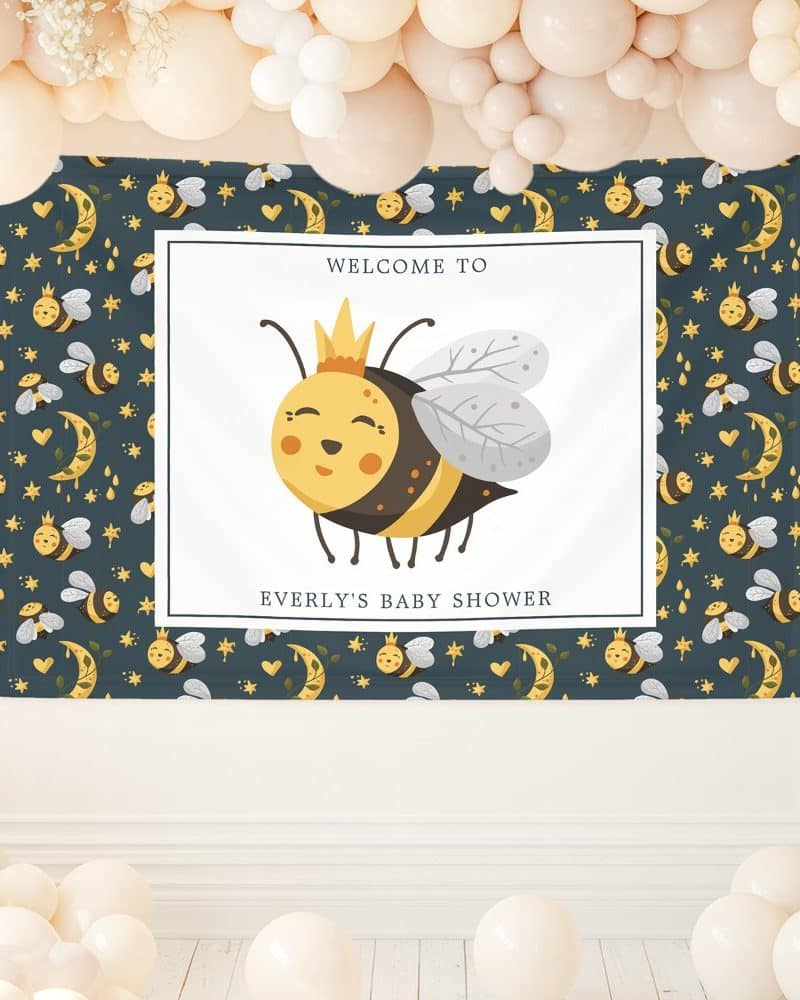A baby shower banner featuring a cartoon bee wearing a crown. The banner has a dark blue background with a repeating pattern of bees, moons, and stars.