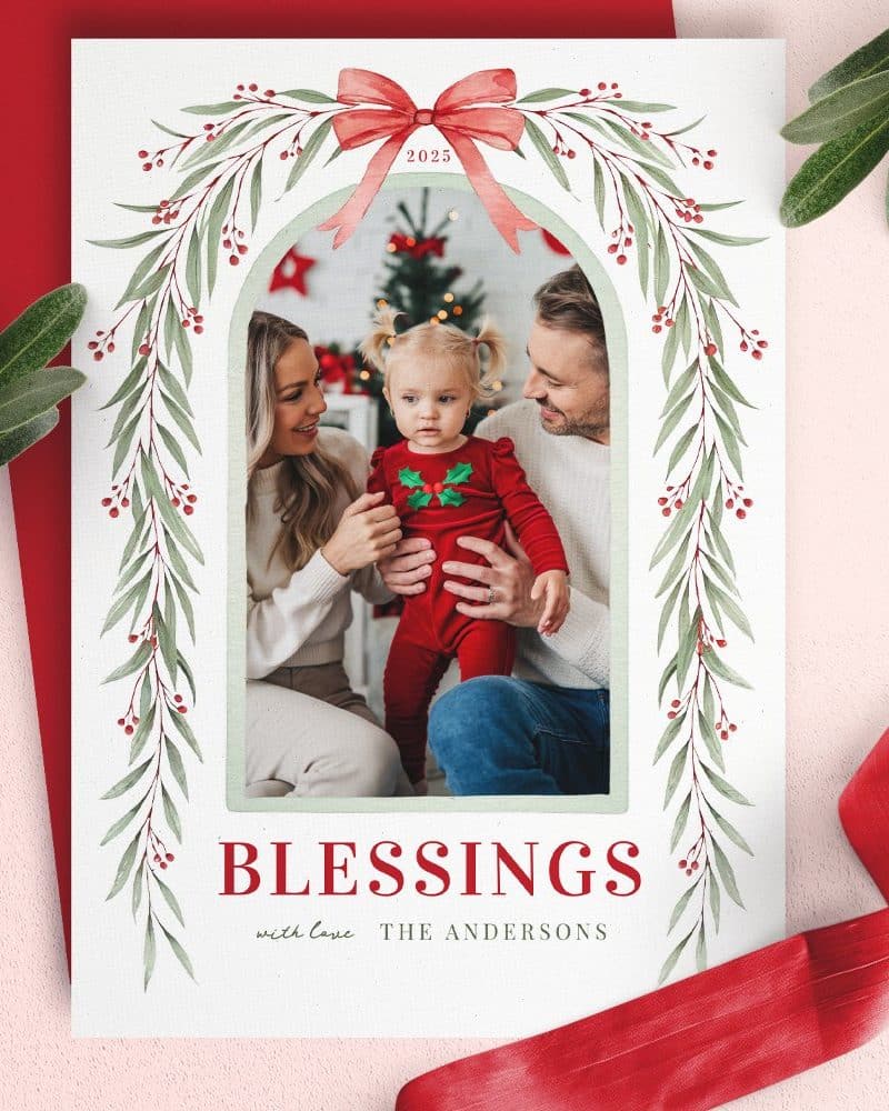 Christmas card featuring a family photo; parents and toddler are smiling. The photo is framed by a garland and a red bow. Text reads "Blessings" and "The Andersons".