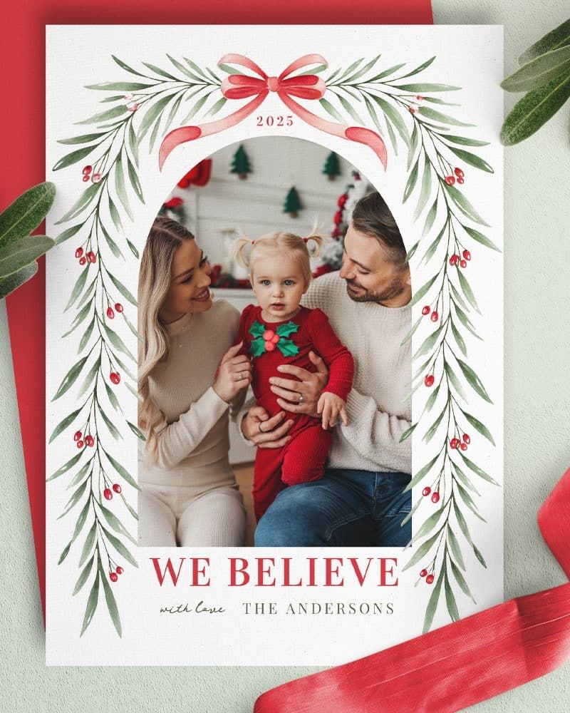 Christmas card featuring a family photo of parents and their toddler framed by a watercolor arch of greenery and berries. The year 2025 and the family's name, The Andersons, are included.