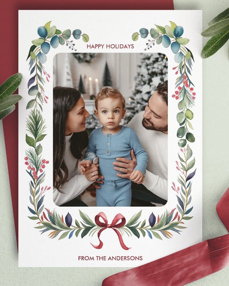 Christmas holiday card featuring a family photo; parents and toddler child are smiling. A watercolor-style floral wreath frames the image.