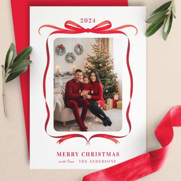 A Christmas Card Featuring A Family Photo Of A Mom, Dad, And Toddler In Red Outfits Sitting In Front Of A Decorated Christmas Tree, Framed By A Red Ribbon Design.