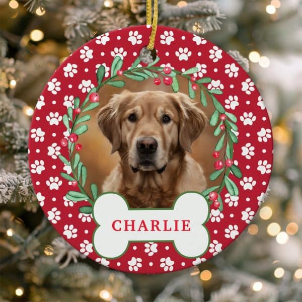Christmas Ornament Featuring A Golden Retriever In A Holly Wreath, With The Name Charlie On A Bone-Shaped Banner. The Ornament Is Red With White Paw Prints.