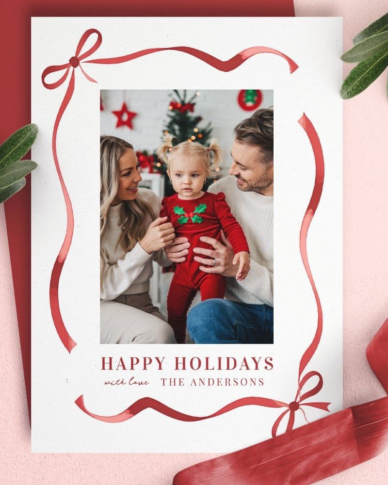 Christmas card featuring a family photo of parents and their toddler daughter. A red ribbon border frames the image.