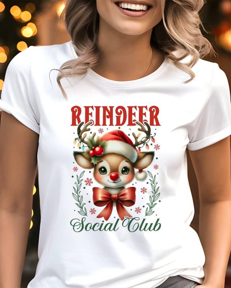 White t-shirt featuring a cute cartoon reindeer wearing a Santa hat and red bow, surrounded by holiday decorations; text reads Reindeer Social Club.