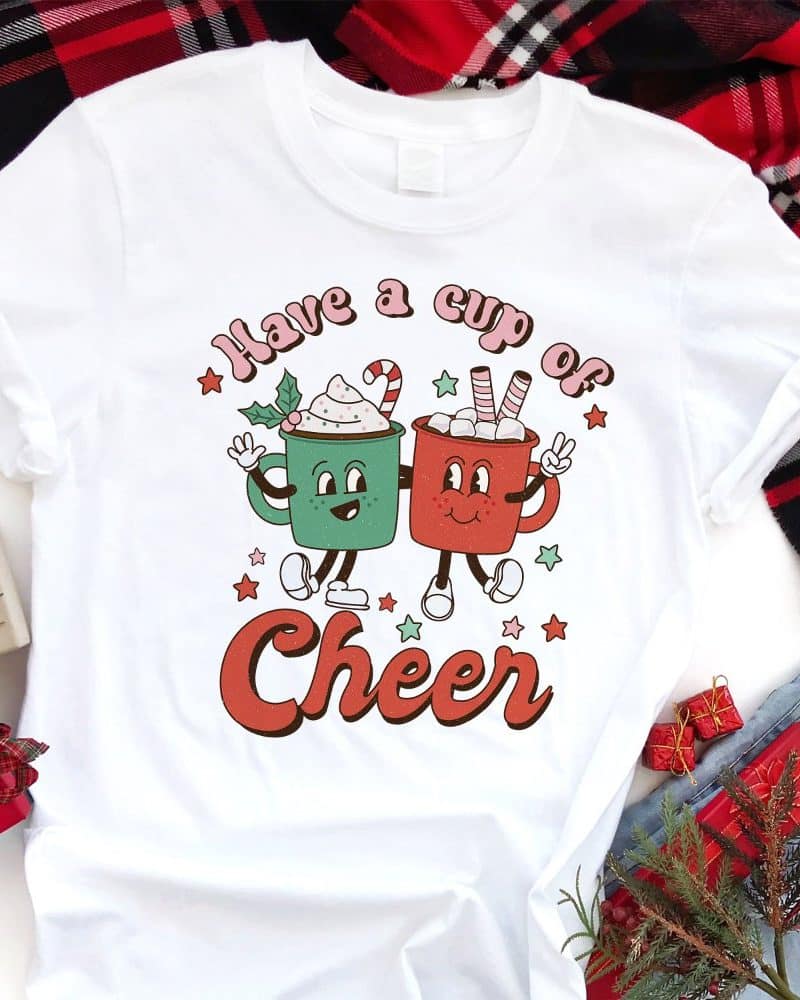 White t-shirt with a Christmas design featuring two cartoon mugs holding hot chocolate, and the text Have a cup of Cheer.
