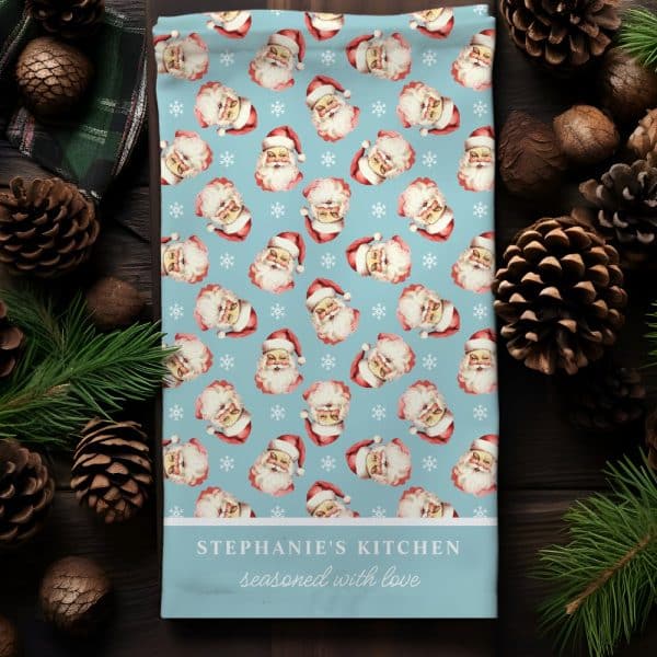 A Light Blue Kitchen Towel With A Repeating Pattern Of Vintage Santa Claus Faces And Snowflakes. The Towel Is Personalized With The Text Stephanie'S Kitchen Seasoned With Love.