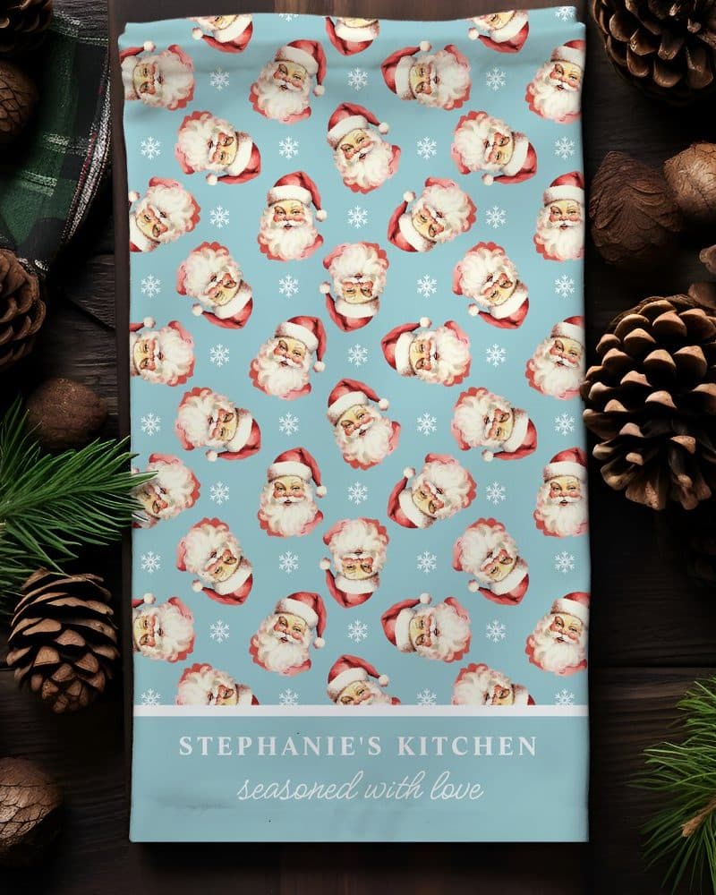 A light blue kitchen towel with a repeating pattern of vintage Santa Claus faces and snowflakes. The towel is personalized with the text Stephanie's Kitchen seasoned with love.