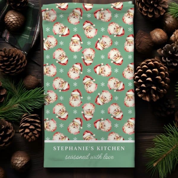 Christmas Tea Towel With A Repeating Pattern Of Santa Claus Faces On A Teal Background. The Towel Is Personalized With The Text Stephanie'S Kitchen Seasoned With Love.