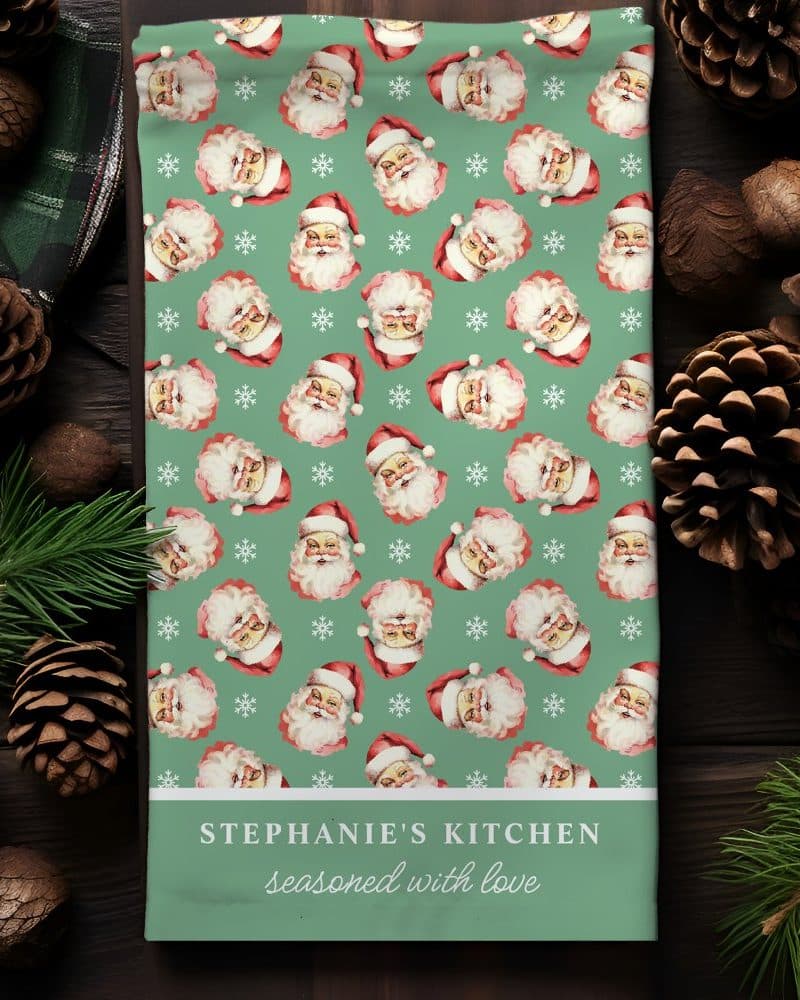 Christmas tea towel with a repeating pattern of Santa Claus faces on a teal background. The towel is personalized with the text Stephanie's Kitchen seasoned with love.