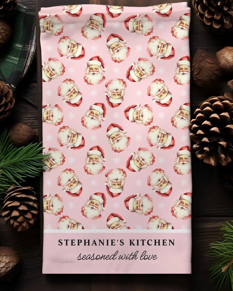 Pink kitchen towel with a repeating pattern of Santa Claus faces and snowflakes. The bottom features personalized text.