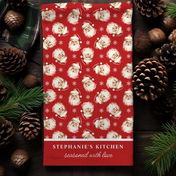 Red Kitchen Towel With A Repeating Pattern Of Santa Claus Faces And Snowflakes; Personalized With The Text Stephanie'S Kitchen Seasoned With Love.