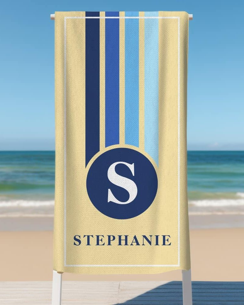 Beige beach towel with blue and light blue vertical stripes, a navy blue circle containing the letter S, and the name Stephanie.