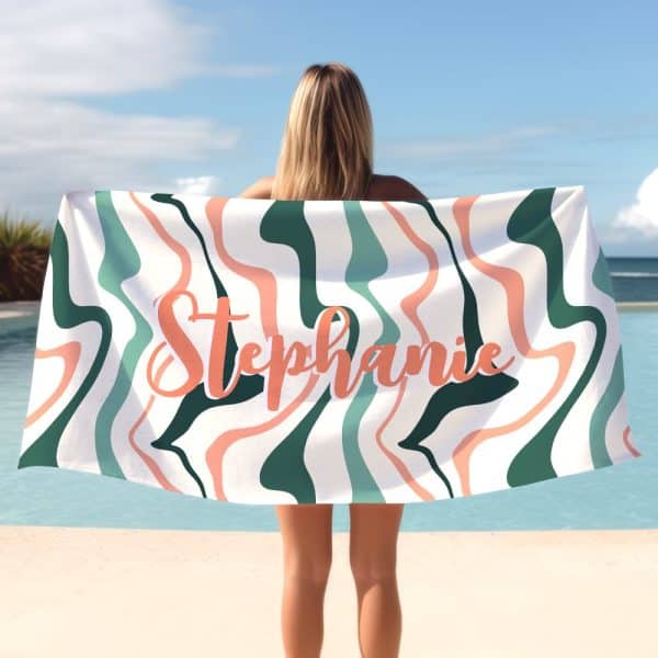 A Woman Holds A Beach Towel Behind Her. The Towel Has A Wavy, Abstract Design In Teal, Peach, And White, With The Name Stephanie Printed In Peach.