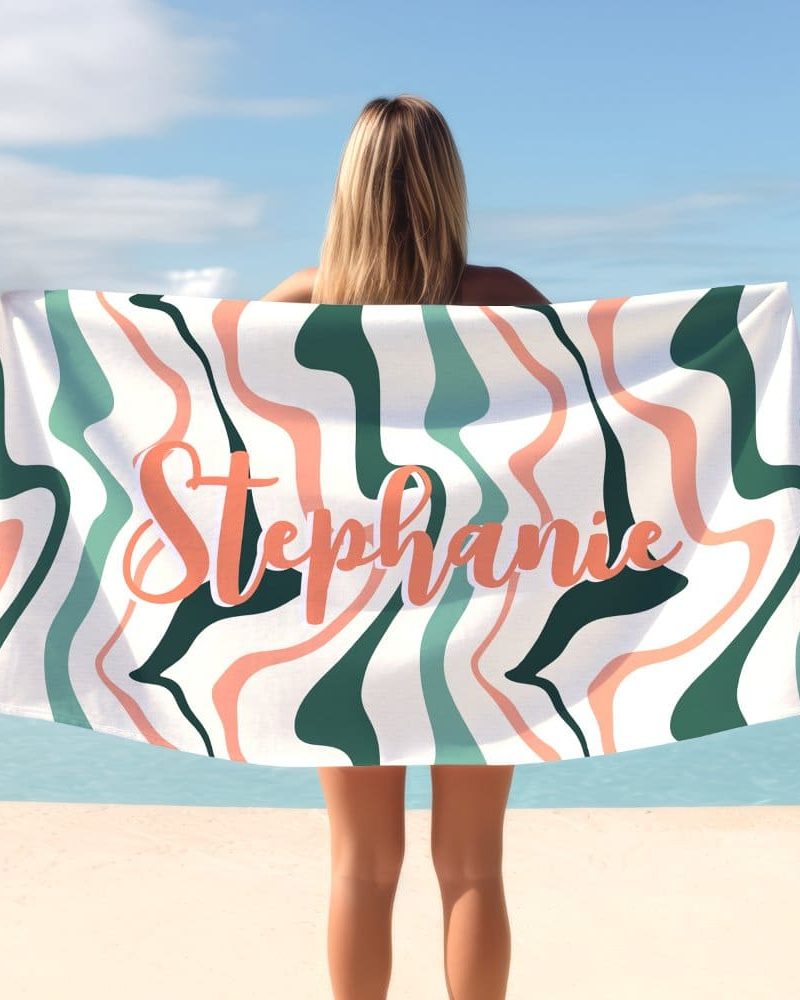 A woman holds a beach towel behind her. The towel has a wavy, abstract design in teal, peach, and white, with the name Stephanie printed in peach.