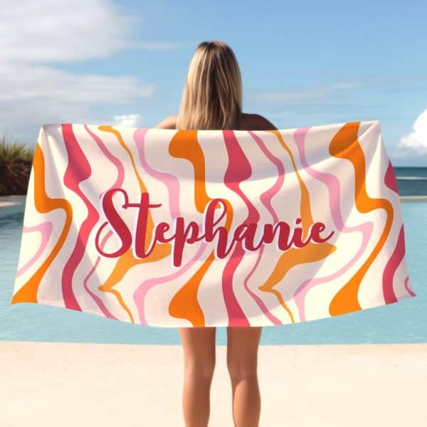 Woman Holding A Personalized Beach Towel With A Swirling Orange, Pink, And Cream Pattern. The Name Stephanie Is Printed On The Towel.