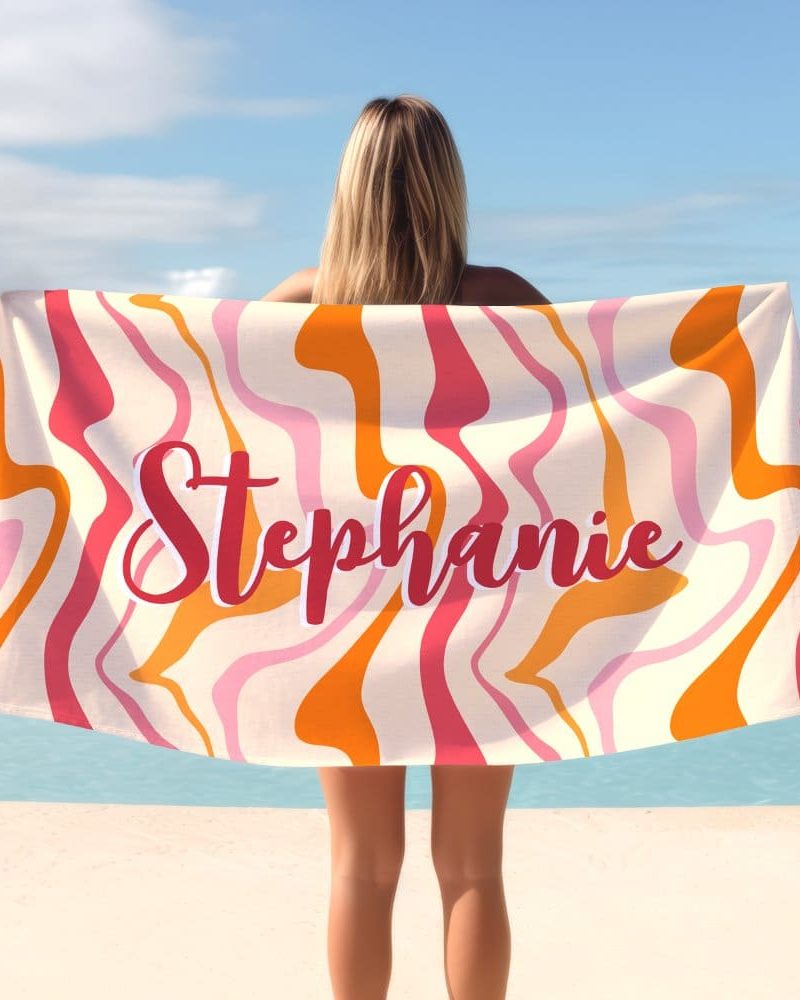 Woman holding a personalized beach towel with a swirling orange, pink, and cream pattern. The name Stephanie is printed on the towel.