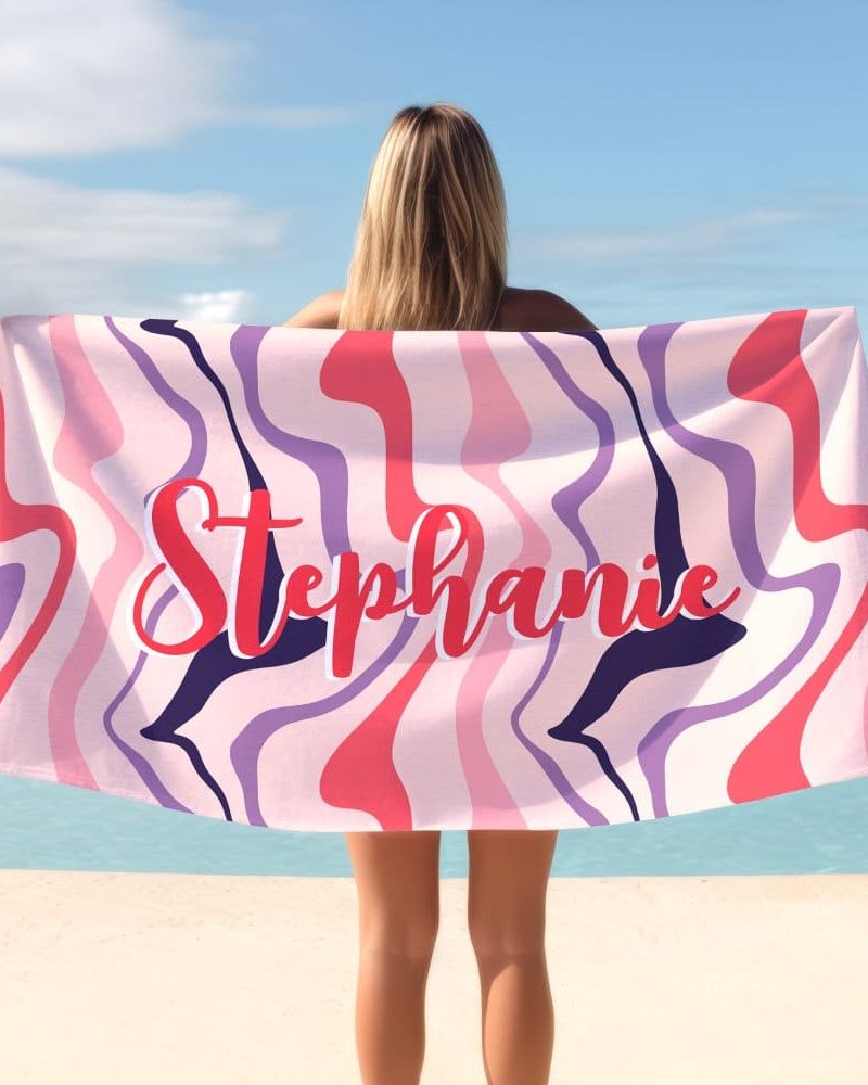 A woman holds a pink, purple, and red beach towel with the name Stephanie written across it in a stylized font.