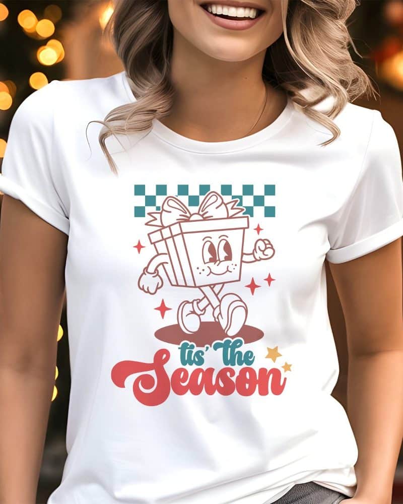 Woman wearing a white t-shirt featuring a retro cartoon gift character walking and the text Tis the Season.