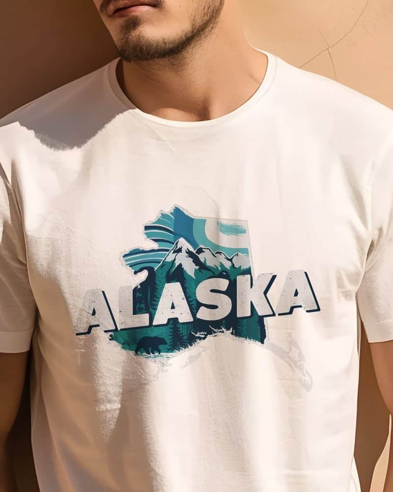 A man is modeling a white t-shirt featuring a teal graphic of Alaska's outline with mountains, trees, and bears.