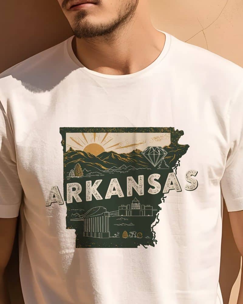Person wearing a white t-shirt with a dark-green graphic of the Arkansas state outline, featuring a sunrise, mountains, and state landmarks.