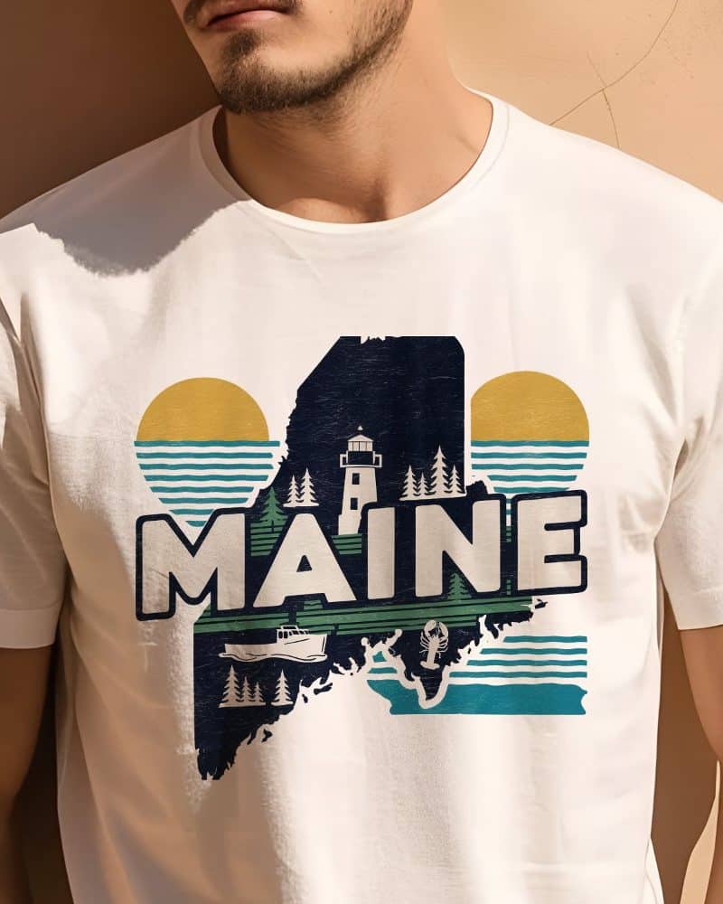 White t-shirt featuring a graphic design of the state of Maine, incorporating a lighthouse, lobster, boat, and stylized sun and waves.