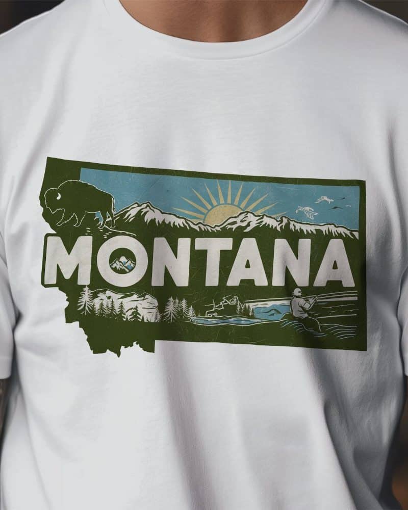White t-shirt with a vintage-style graphic of the state of Montana. The design includes mountains, a bison, a sun, and a person fishing.