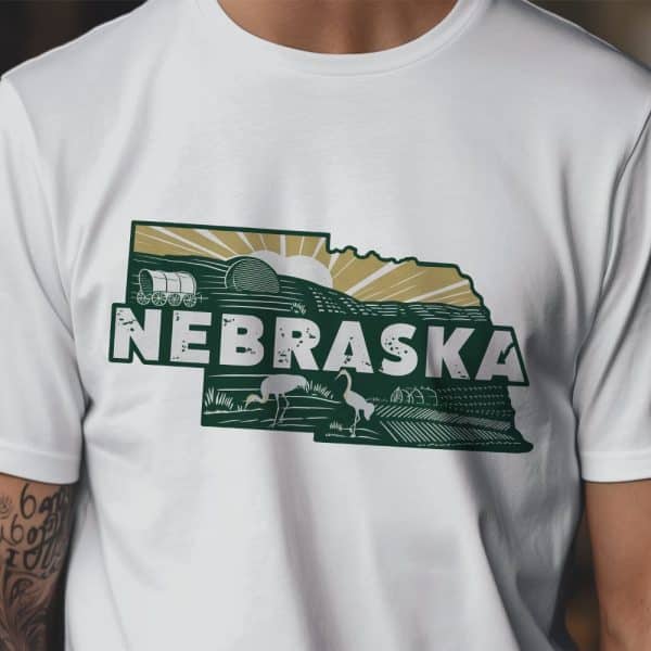 White T-Shirt With A Dark Green Nebraska-Shaped Graphic Featuring A Sunrise, Wagon, Birds, And Farmland.