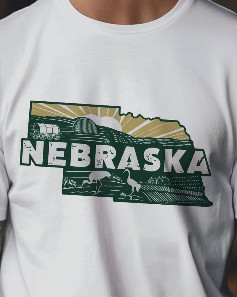 White t-shirt with a dark green Nebraska-shaped graphic featuring a sunrise, wagon, birds, and farmland.