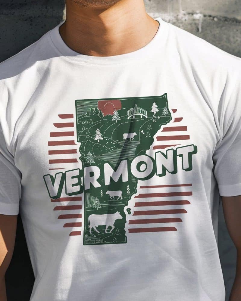 White t-shirt featuring a green outline of Vermont with illustrations of cows and landscape, and the word Vermont in bold text.