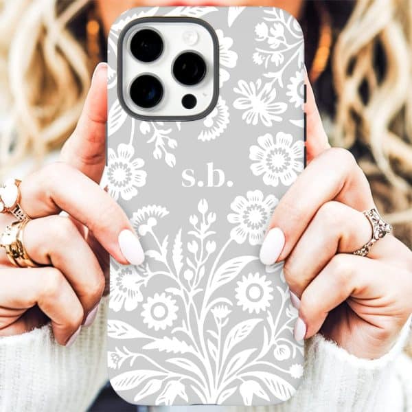 A Woman'S Hands Holding A Light Gray Iphone Case With A White Floral Pattern And The Initials S.b.