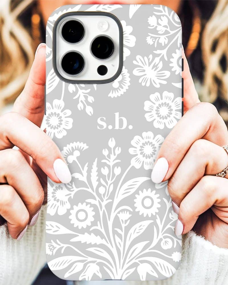 A woman's hands holding a light gray iPhone case with a white floral pattern and the initials s.b.