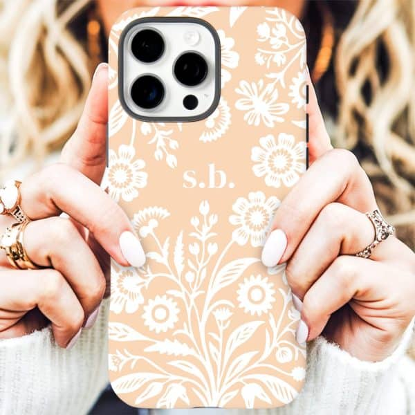 Close-Up Of Hands Holding A Peach-Colored Iphone Case With A White Floral Pattern And Initials Sb.