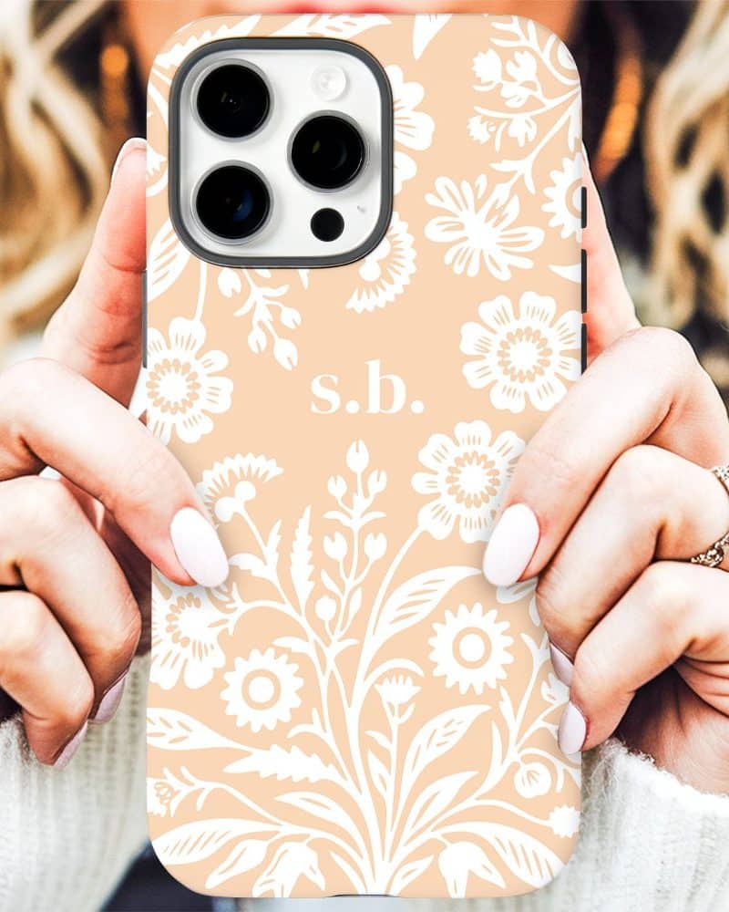 Close-up of hands holding a peach-colored iPhone case with a white floral pattern and initials SB.