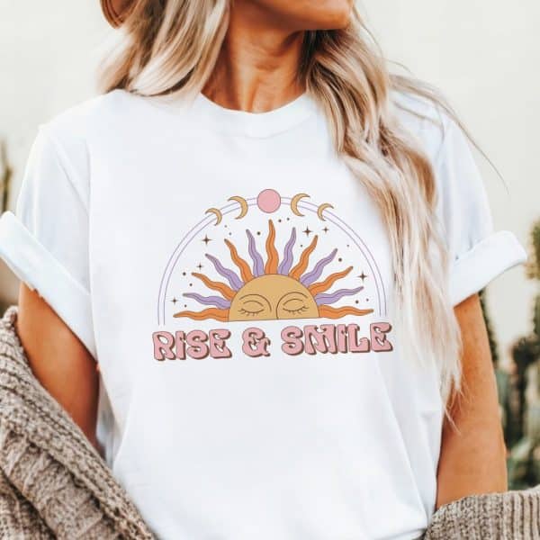 White T-Shirt With A Stylized Sun, Moon Phases, And Stars; Text Reads Rise &Amp; Smile.