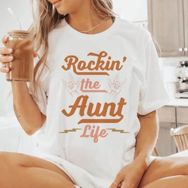 Woman Wearing A White T-Shirt That Says Rockin' The Aunt Life In A Retro Style Font.