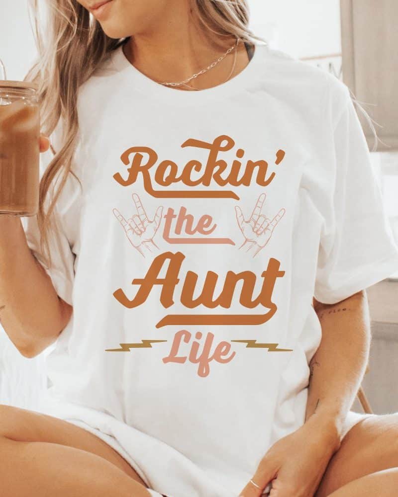Woman wearing a white t-shirt that says Rockin' the Aunt Life in a retro style font.