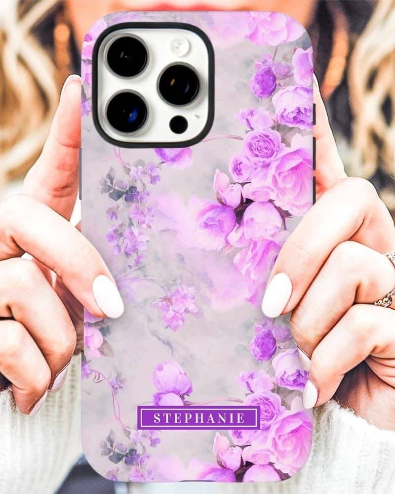 A woman's hands hold up an iPhone case featuring a watercolor design of light purple roses on a gray marble background. The name Stephanie is printed on the case.