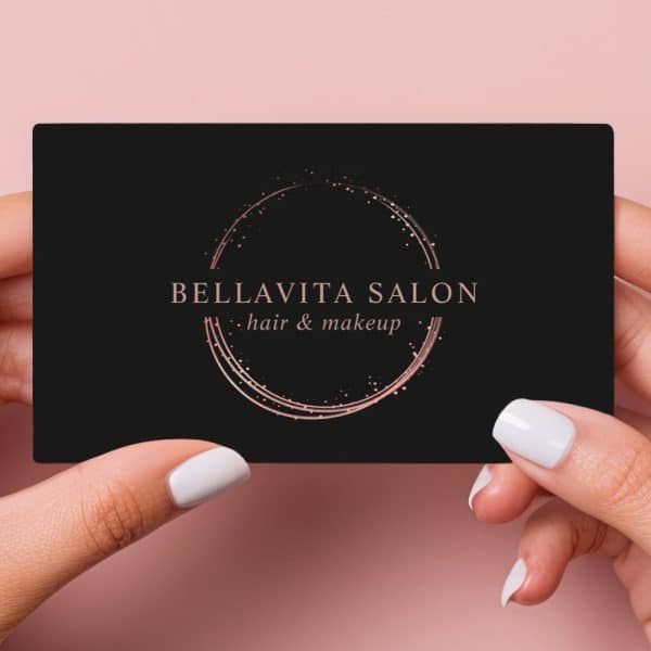Two Hands Hold Up A Black Business Card With Rose Gold Lettering. The Card Advertises Bellavita Salon, Hair And Makeup.