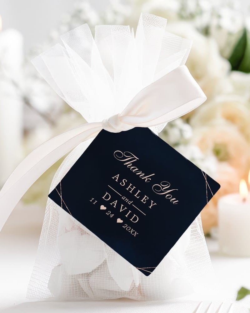A white tulle bag tied with a cream ribbon and containing white petals, with a navy blue thank you tag that says Ashley and David's names and wedding date.