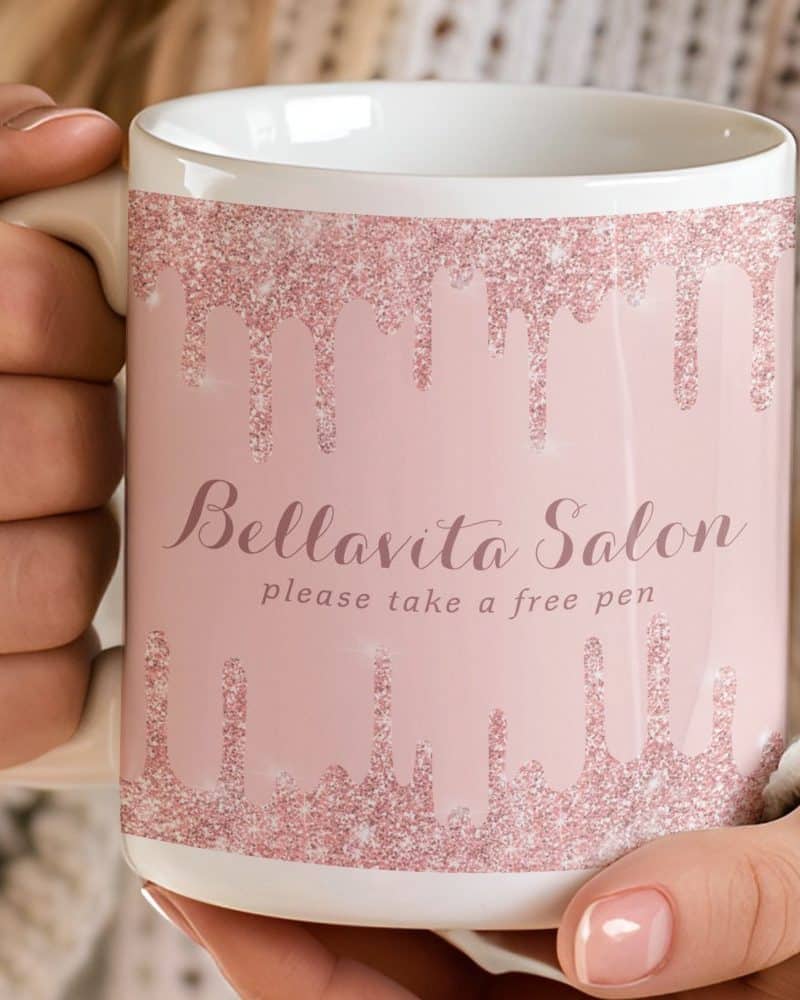 A person holding a pink mug with rose gold glitter dripping down the sides. The mug says Bellavita Salon and Please take a free pen.