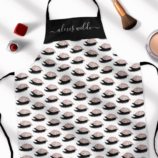 A Personalized Apron With A Repeating Pattern Of Glitter Eyelashes. The Name Alexis Wilde Is On The Black Top Section.