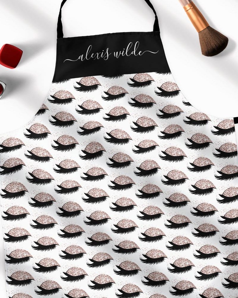 A personalized apron with a repeating pattern of glitter eyelashes. The name Alexis Wilde is on the black top section.
