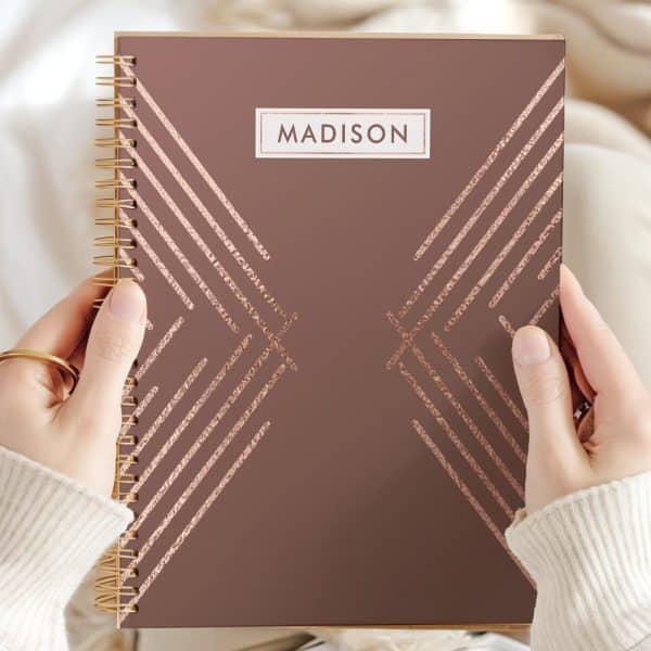 Close-Up Of Hands Holding A Mauve Spiral-Bound Notebook With Rose Gold Geometric Lines And The Name Madison On The Cover.