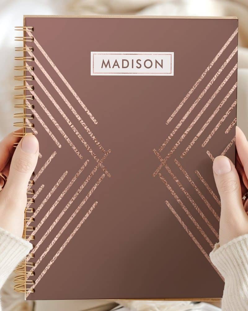 Close-up of hands holding a mauve spiral-bound notebook with rose gold geometric lines and the name Madison on the cover.