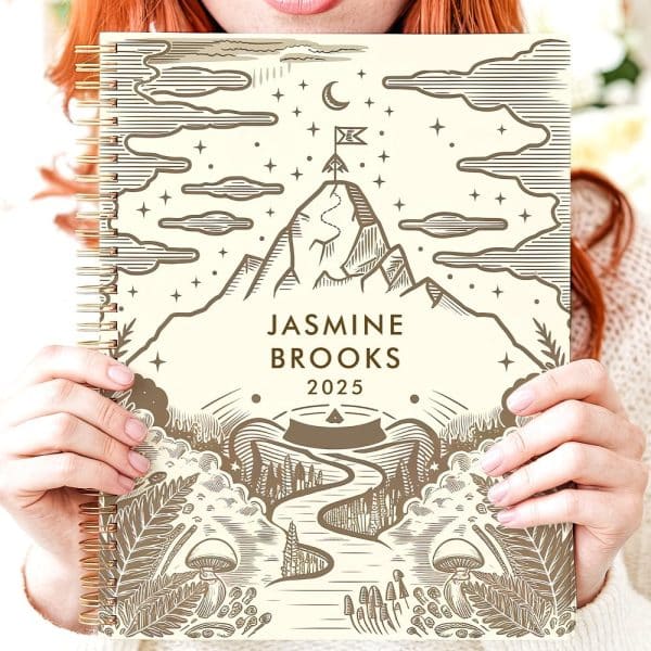 Person Holding A 2025 Planner Featuring A Mountain Scene With A Winding Path.