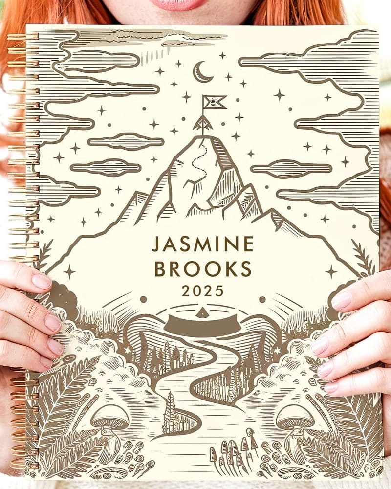 Person holding a 2025 planner featuring a mountain scene with a winding path.
