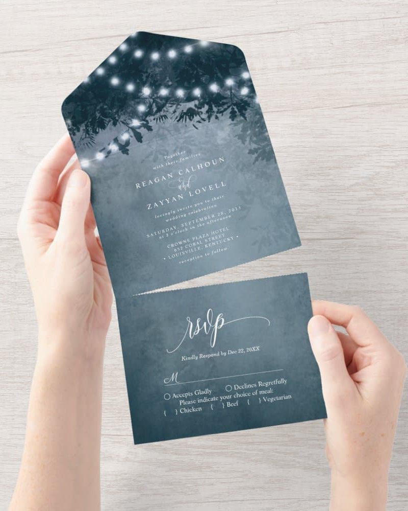 A blue wedding invitation with string lights and tree branches in the background. The invitation includes the couple's names and wedding details, along with an RSVP card.