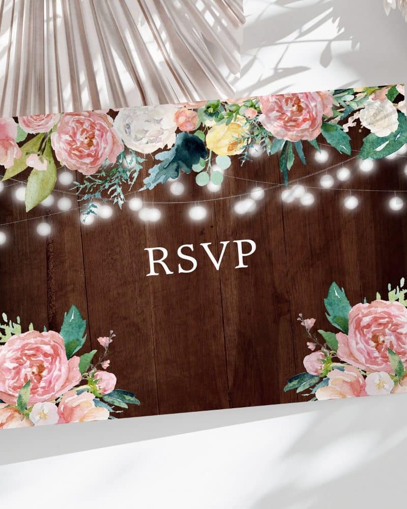 RSVP card with pink and peach floral watercolor design and string lights on a dark wood background.